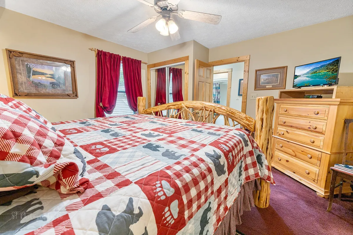 A warm and inviting king bedroom featuring a rustic log bed frame dressed in a buffalo check quilt, complemented by red curtains and a wooden dresser. The bedroom exudes a cozy, tranquil atmosphere, and includes decorative artwork on the walls. The presence of a TV visible from the bed aligns with the in-room entertainment described in the section.