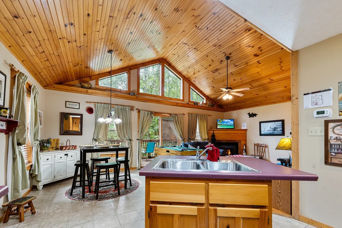A warm and welcoming open-plan area featuring large windows that offer glimpses of the outdoors, a spacious dining table for group meals, and a kitchen island perfect for social gatherings. The wooden ceiling and overall rustic charm hint at the cabin's natural surroundings and its connection to the outdoors.