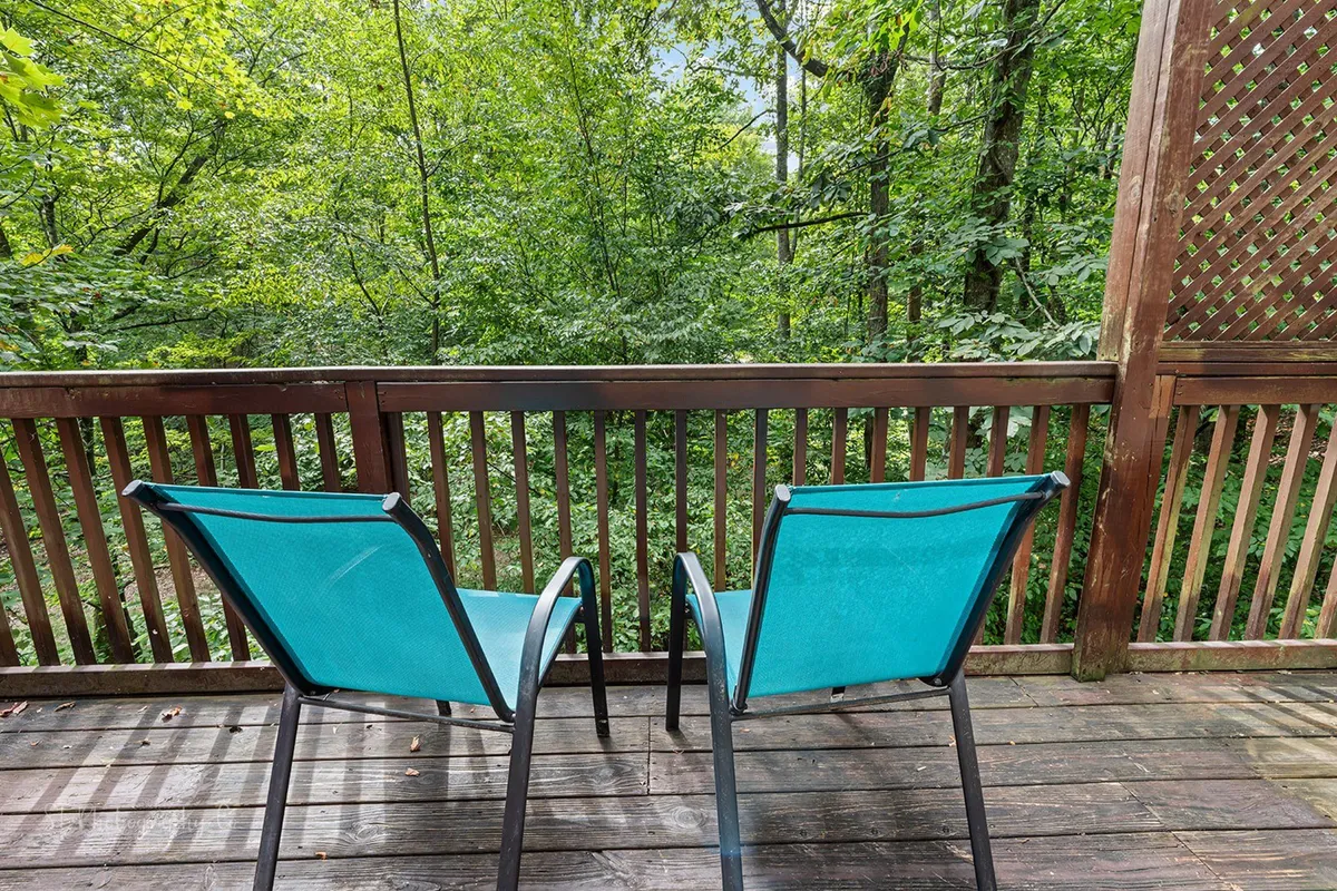 A tranquil exterior view featuring two turquoise chairs situated on a wooden deck, enclosed by a railing and surrounded by lush greenery. The setup suggests a private, serene spot perfect for relaxing, reading, or gazing at the natural landscape.