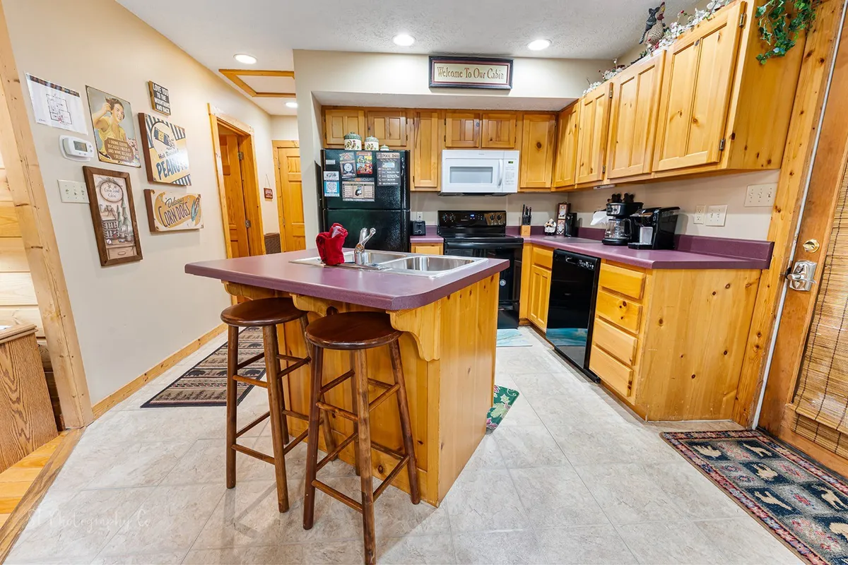 A warm, inviting kitchen with a central island, wood cabinetry, and black appliances. Decorative signs add personality to the space, suggesting a homey atmosphere perfect for family gatherings or casual dining. The layout highlights modern appliances and ample counter space, with plenty of cookware and dishes on display.