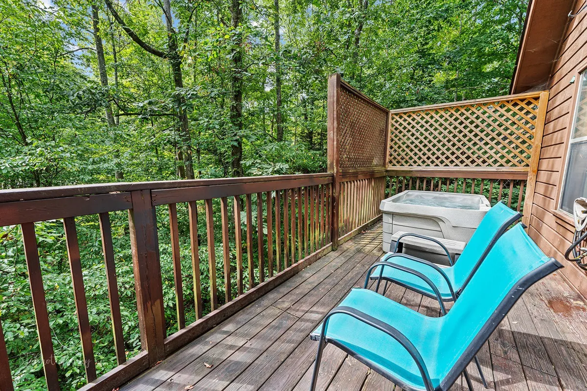 A peaceful deck bordered by trees, featuring a cozy hot tub and bright blue chairs for outdoor seating. The setting is perfect for relaxation, with options to lounge, read, or enjoy the scenic wooded surroundings.