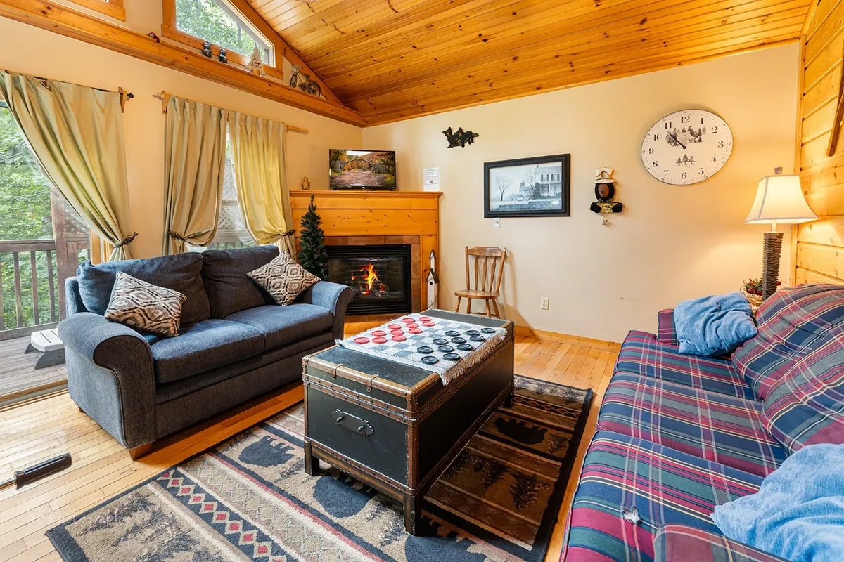 A warm interior scene featuring a comfortable sofa, a central fireplace, a coffee table, and large windows offering wooded views. The room is styled for comfort, perfect for cozy evenings and intimate gatherings.