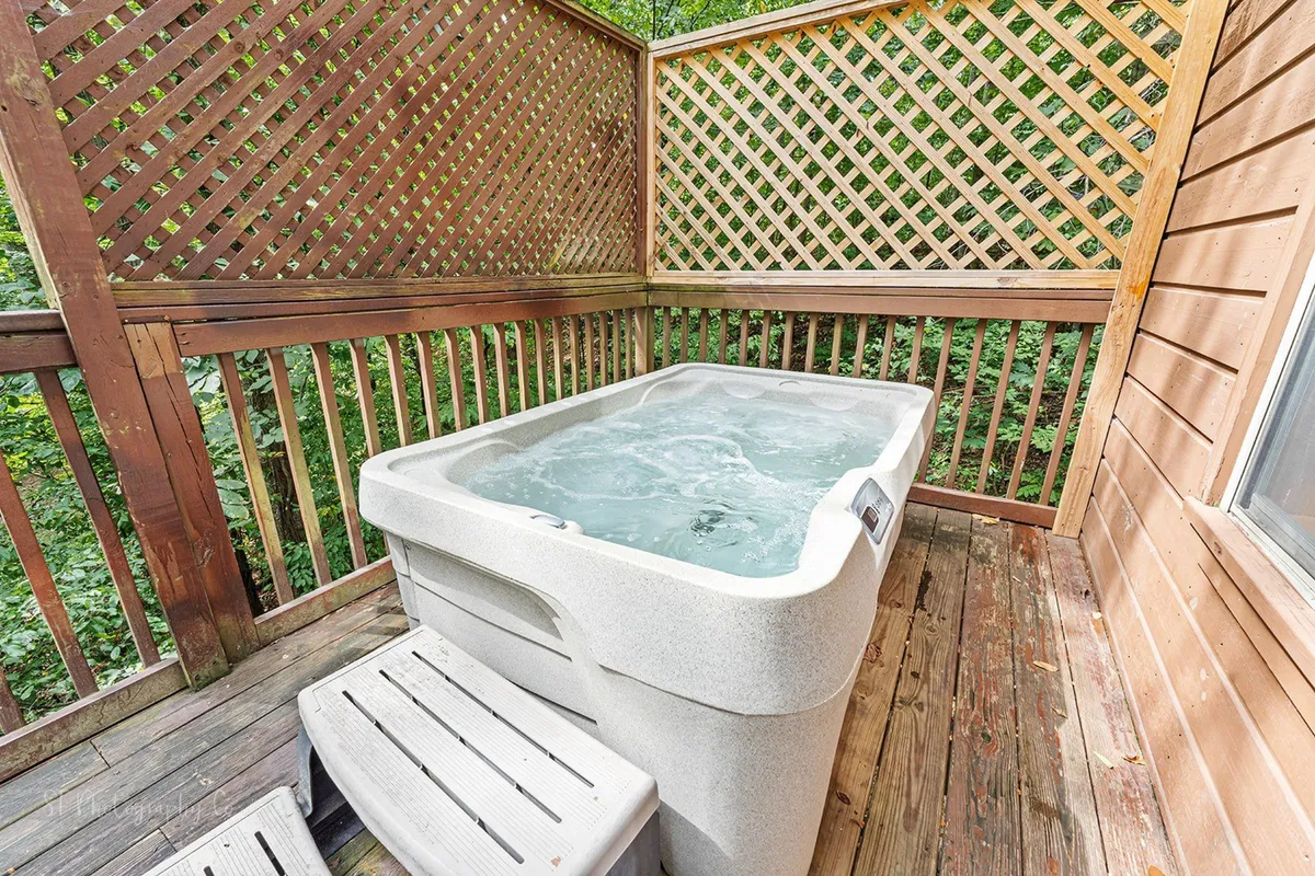 A serene wooden deck surrounded by a lattice fence, featuring a bubbling hot tub with space for four. The deck is nestled among trees, creating a tranquil, private oasis perfect for unwinding. The scene suggests both relaxation in the hot tub and enjoying the crisp mountain air, with stargazing or morning coffee options.