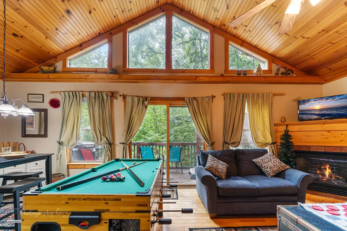 An inviting, multifunctional living area displaying a pool table, fireplace, sofa, dining table, and expansive windows letting in natural light. The scene blends recreational opportunities and comfort in one cohesive space.