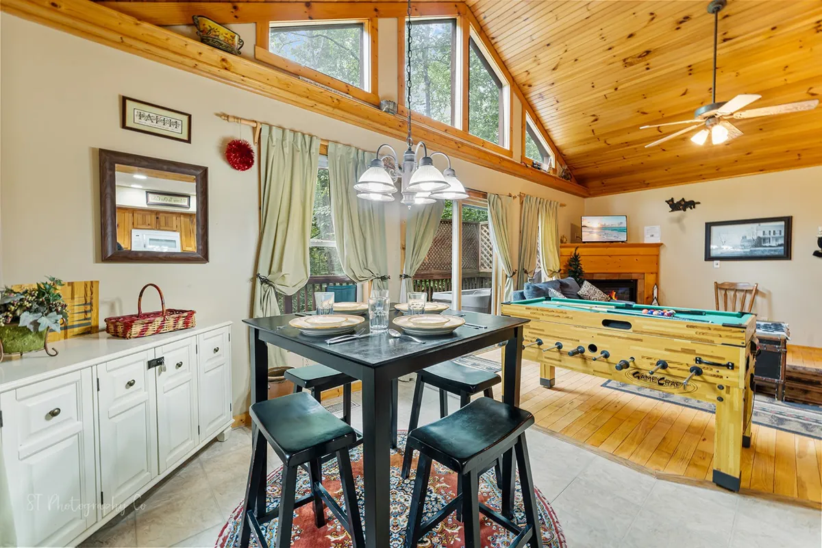 A bright, open interior space featuring a dining table and a pool table, with large windows that offer a view of the surrounding woods. The wooden ceiling and expansive glazing create a strong visual connection between the indoors and the outdoor environment, suggesting the cabin’s proximity to natural adventures.