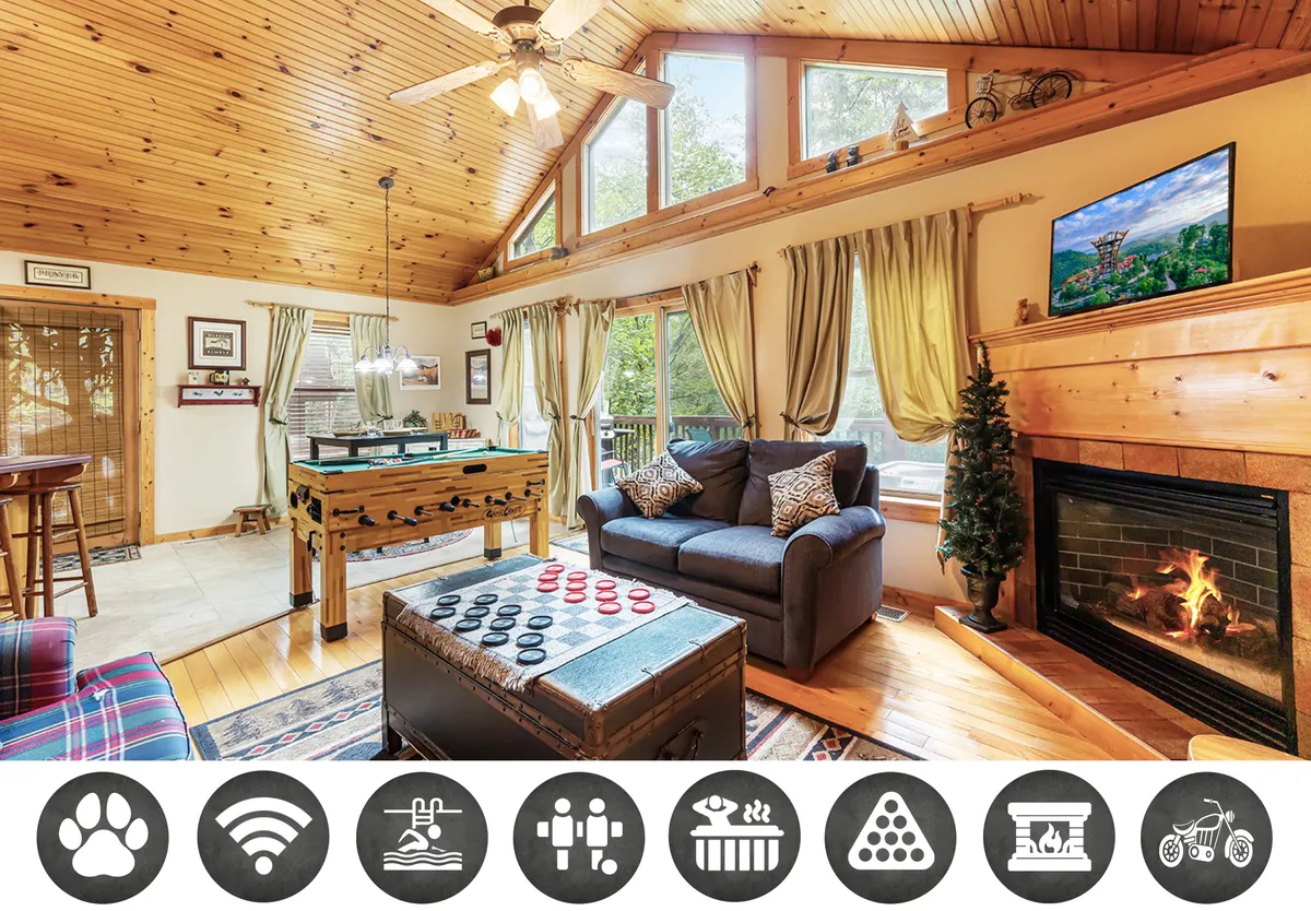 The image shows a cozy, open-plan cabin interior with a plush sofa facing a stone fireplace, a foosball table ready for friendly competition, and expansive windows that frame lush Smoky Mountain views. The warm wood finishes and soft lighting create an inviting atmosphere, perfect for relaxation and togetherness.