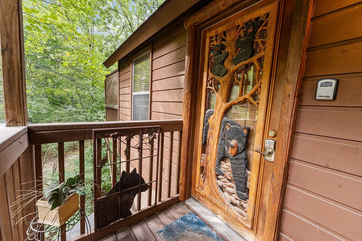 A close-up of the cabin’s decorative wooden front door featuring bear carvings, with rustic wooden railings and lush greenery visible. The artistic entryway is both charming and memorable, setting the stage for a romantic getaway.
