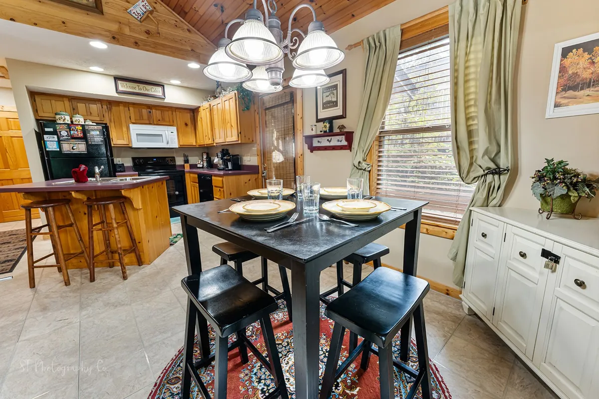 A cozy dining area featuring a wooden table, elegant chandelier, window with curtains, and a decorative plant. The setup is ideal for gathering around for family meals, casual dining, or entertaining guests in a warm, homey environment.