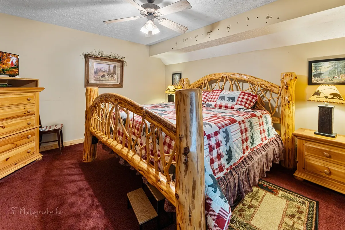 A close-up view of a cozy king bedroom with a rustic log bed, plaid bedding, a wooden dresser, and a television facing the bed. The intimate setting emphasizes comfort and relaxation, ideal for winding down with a movie or a good book.