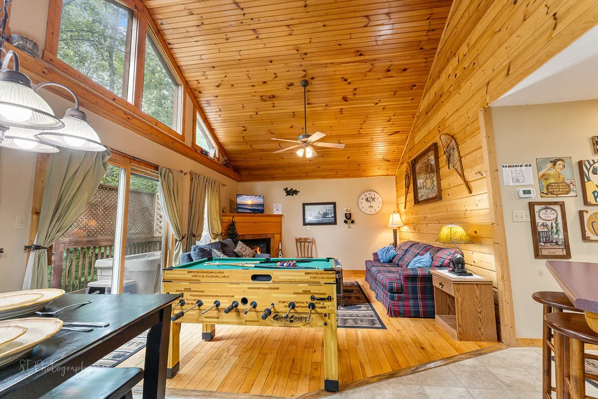 A bright, family-friendly living space featuring a foosball table, a cozy plaid sofa, wooden ceilings, and large windows. The room exudes a welcoming atmosphere perfect for indoor games, socializing, and relaxing with loved ones after a day exploring local attractions.